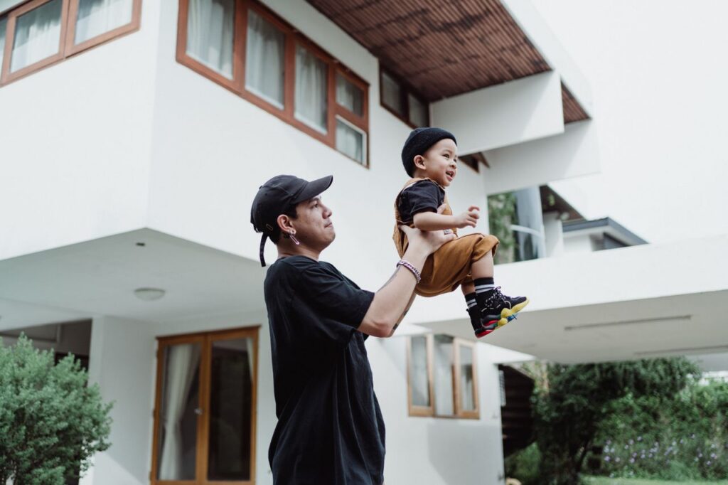 A father lifts his smiling son in front of a contemporary house, showcasing a joyful family moment.
